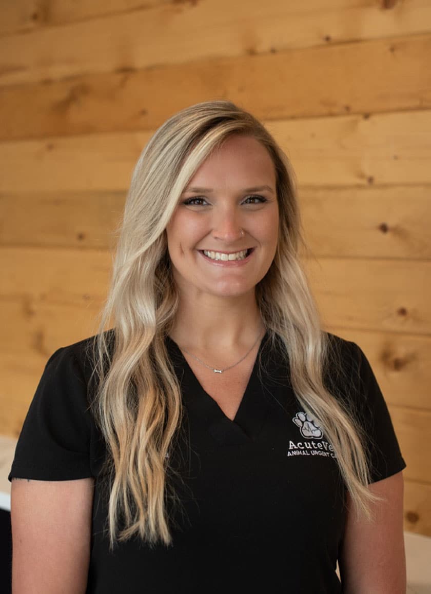 A smiling person with long blonde hair stands against a wooden background. They are wearing a black shirt with a logo and text that reads "Acute Care Animal Urgent Care," showcasing their pride as a veterinarian.