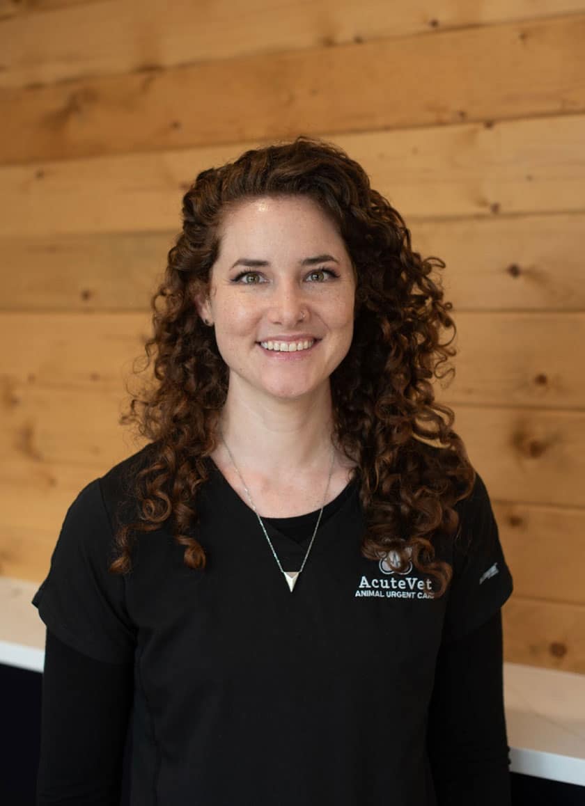 A woman with long curly hair, wearing a black shirt with the logo "AcuteVet Animal Urgent Care," stands in front of a wood-paneled wall. She is smiling and has a necklace with a small, triangular pendant. The veterinarian's warm demeanor reflects her dedication to caring for animals.