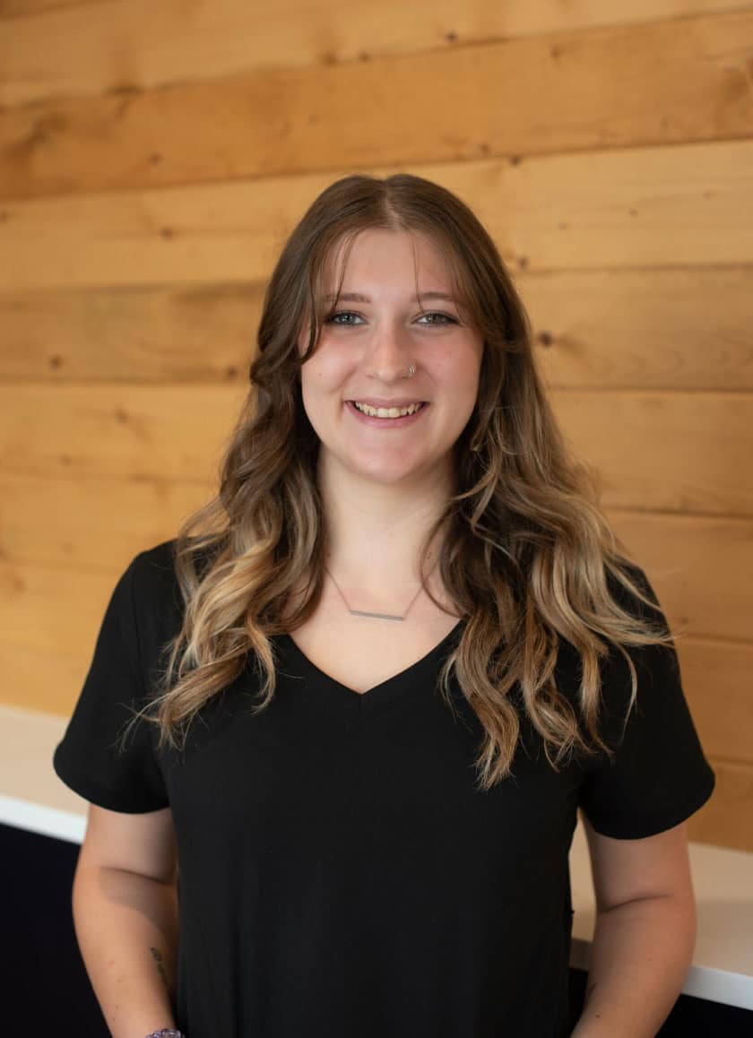 Woman with long, wavy, brunette hair and subtle blonde highlights, wearing a black V-neck shirt and a necklace, smiling as she stands in front of a wooden-paneled wall; she is a compassionate veterinarian dedicated to animal care.