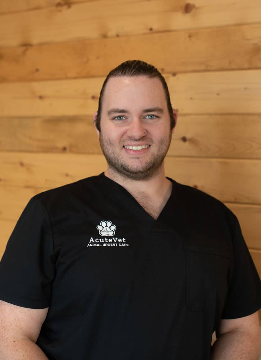 A man with a light complexion and long, tied-back hair is smiling at the camera. He is wearing a black scrub top with the logo and text "Acutevet Animal Urgent Care" on it, highlighting his role as a dedicated veterinarian. The background is a wood-paneled wall.