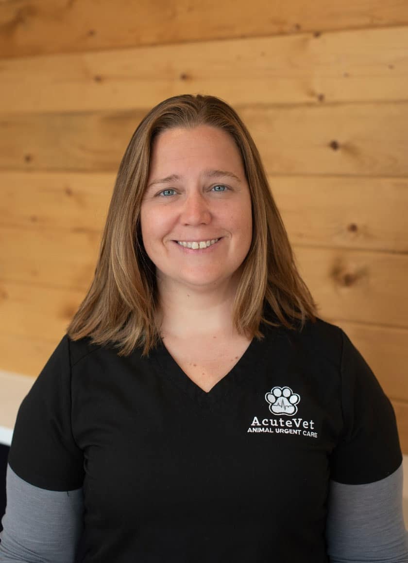 A woman with shoulder-length brown hair, wearing a black V-neck shirt with the "AcuteVet Animal Urgent Care" logo, smiles at the camera. The background features a wooden wall with a natural finish, perfectly capturing her as a compassionate veterinarian dedicated to her work.
