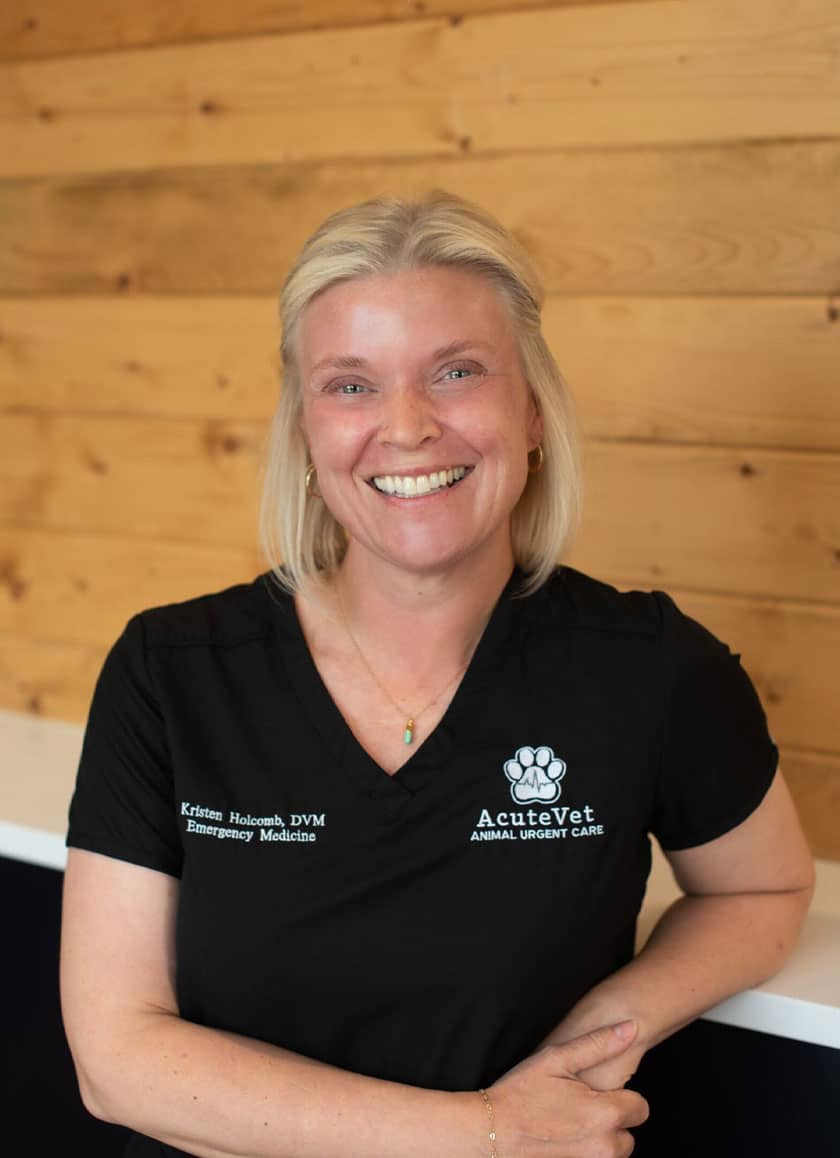 A person with short blonde hair smiles at the camera, wearing a black scrub top with the words "AcuteVet Animal Urgent Care" and "Emergency Medicine" embroidered on it. The veterinarian stands in front of a wooden wall, exuding confidence and warmth.
