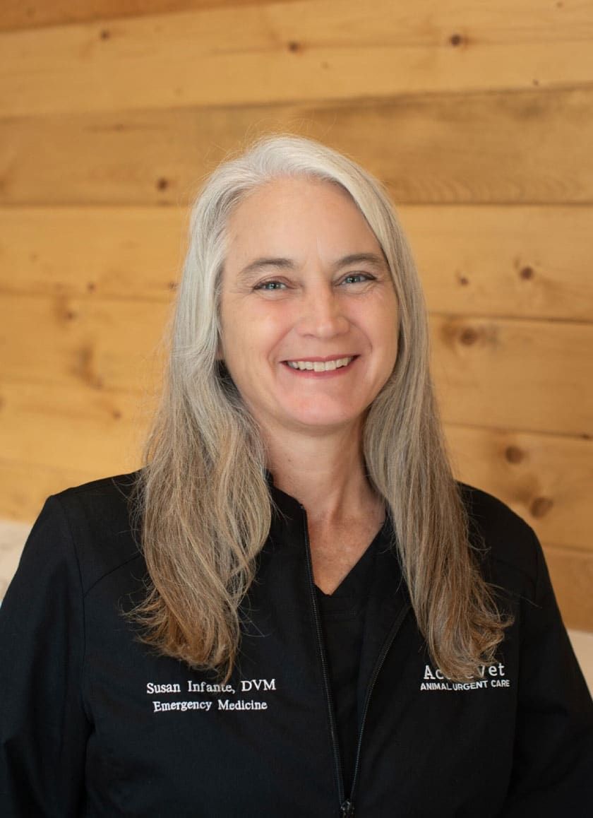 A woman with long gray hair smiling. She is wearing a black jacket with text on it that reads "Susan Infante, DVM, Emergency Medicine" and "AcreVet, Animal Urgent Care." The background consists of wooden planks, highlighting her role as a dedicated veterinarian.