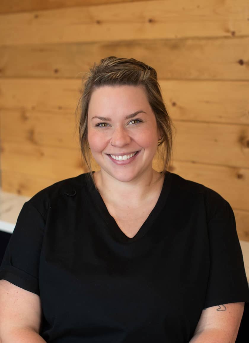 A woman with light brown hair tied back is smiling at the camera. She is a veterinarian wearing a black shirt and standing in front of a wooden wall.
