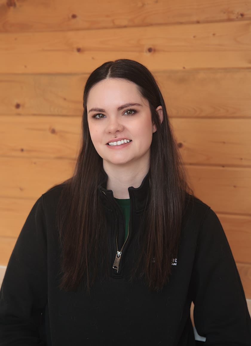 A young woman with long brown hair, wearing a black zip-up jacket, smiles while sitting in front of a wooden wall background.