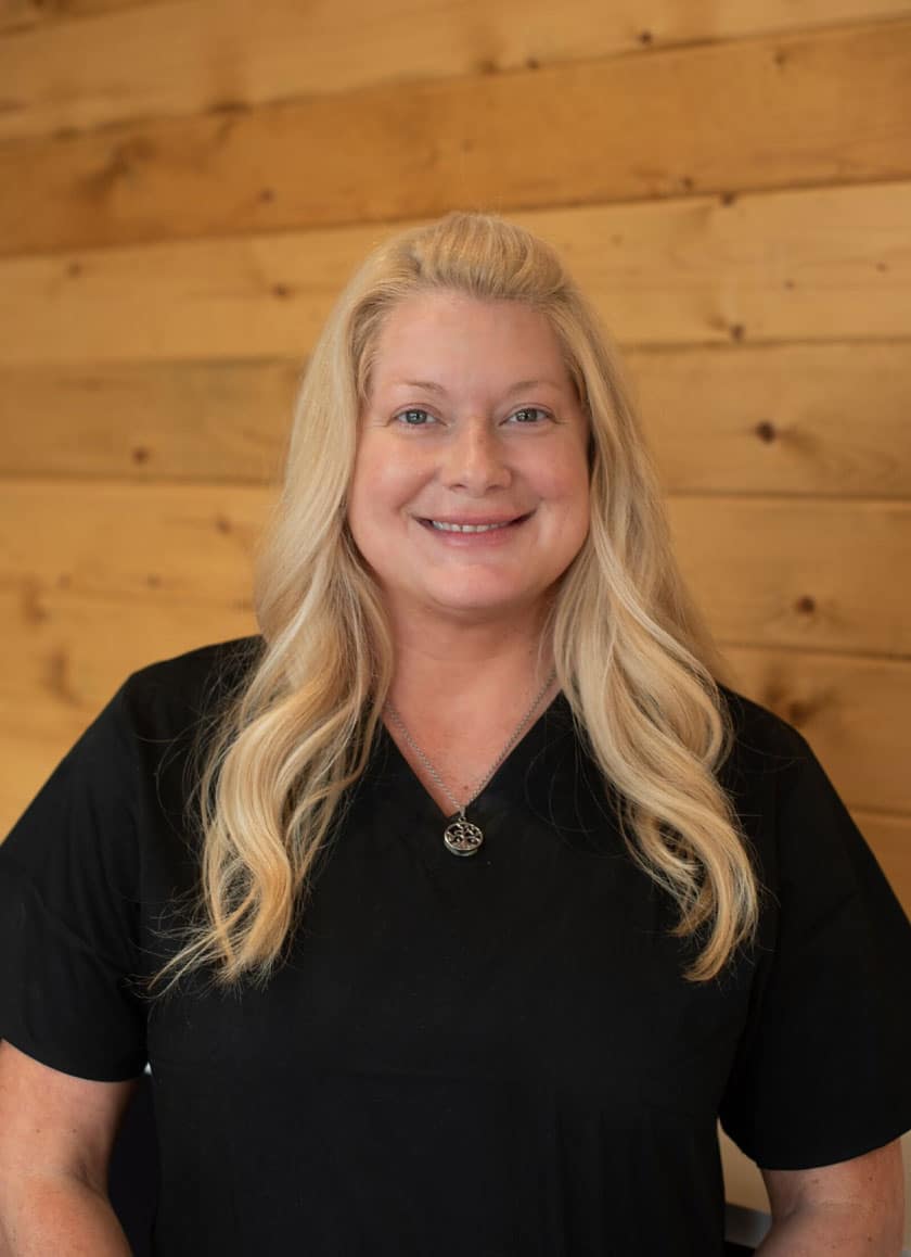 A woman with long blonde hair and wearing a black top is smiling at the camera. She is a veterinarian standing in front of a wooden panel wall.