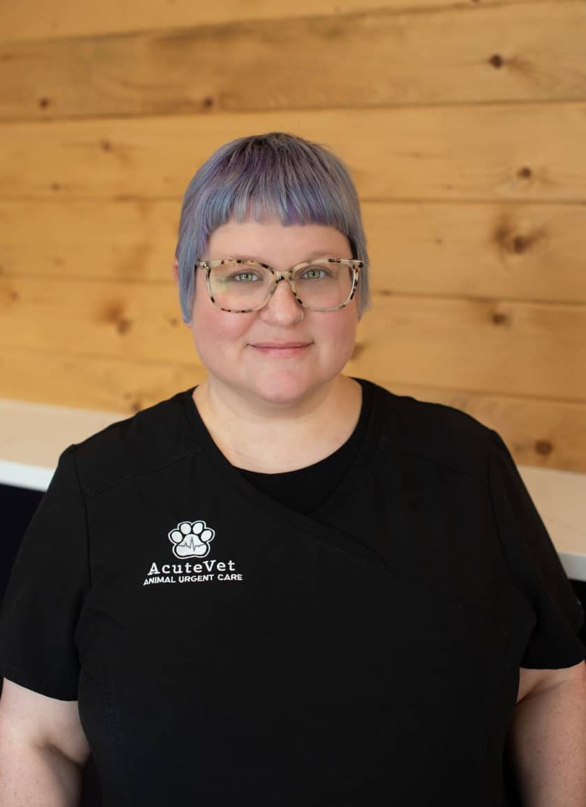 A veterinarian with short lavender hair and glasses poses for a photo against a wooden backdrop. They are wearing a black shirt with the "Acute Vet Animal Urgent Care" logo on the chest.