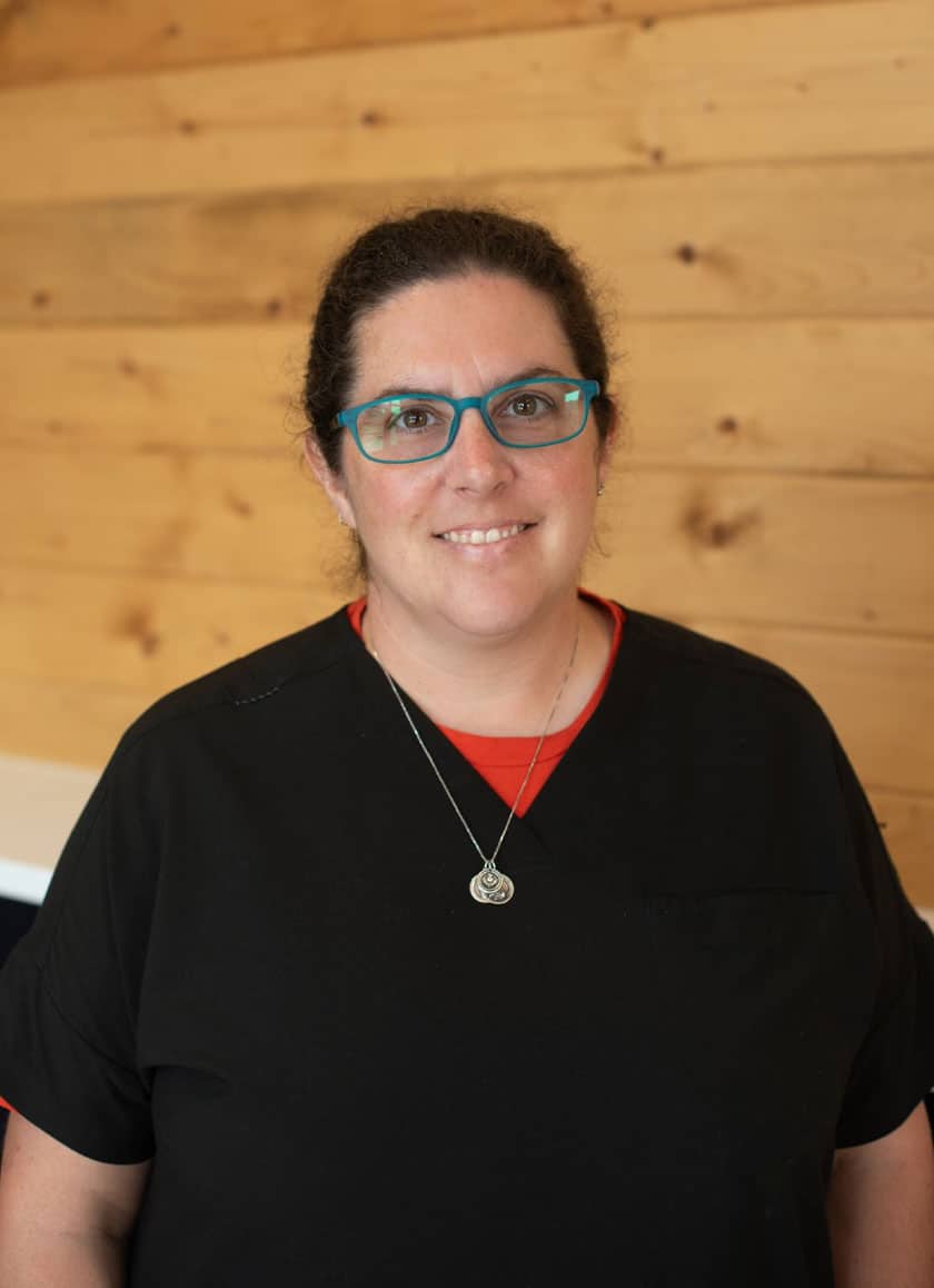 A veterinarian with glasses, wearing a black shirt and a necklace, smiles at the camera. They are standing in front of a wooden wall.