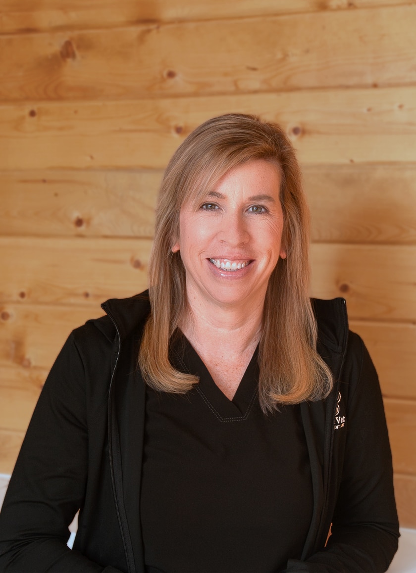 A woman with long blonde hair wearing a black shirt and jacket smiles while seated in front of a wooden wall.