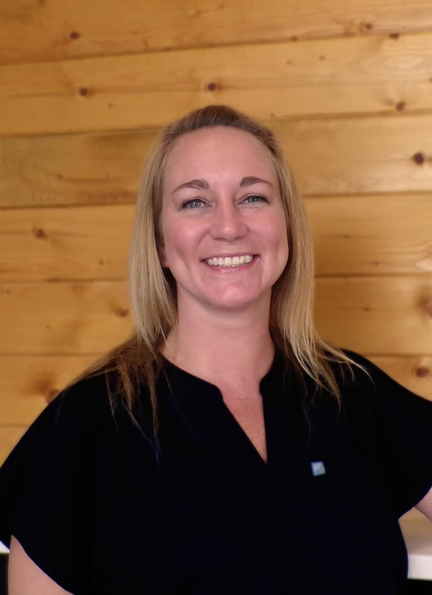 A woman with blonde hair wearing a black top smiles in front of a wooden wall background.