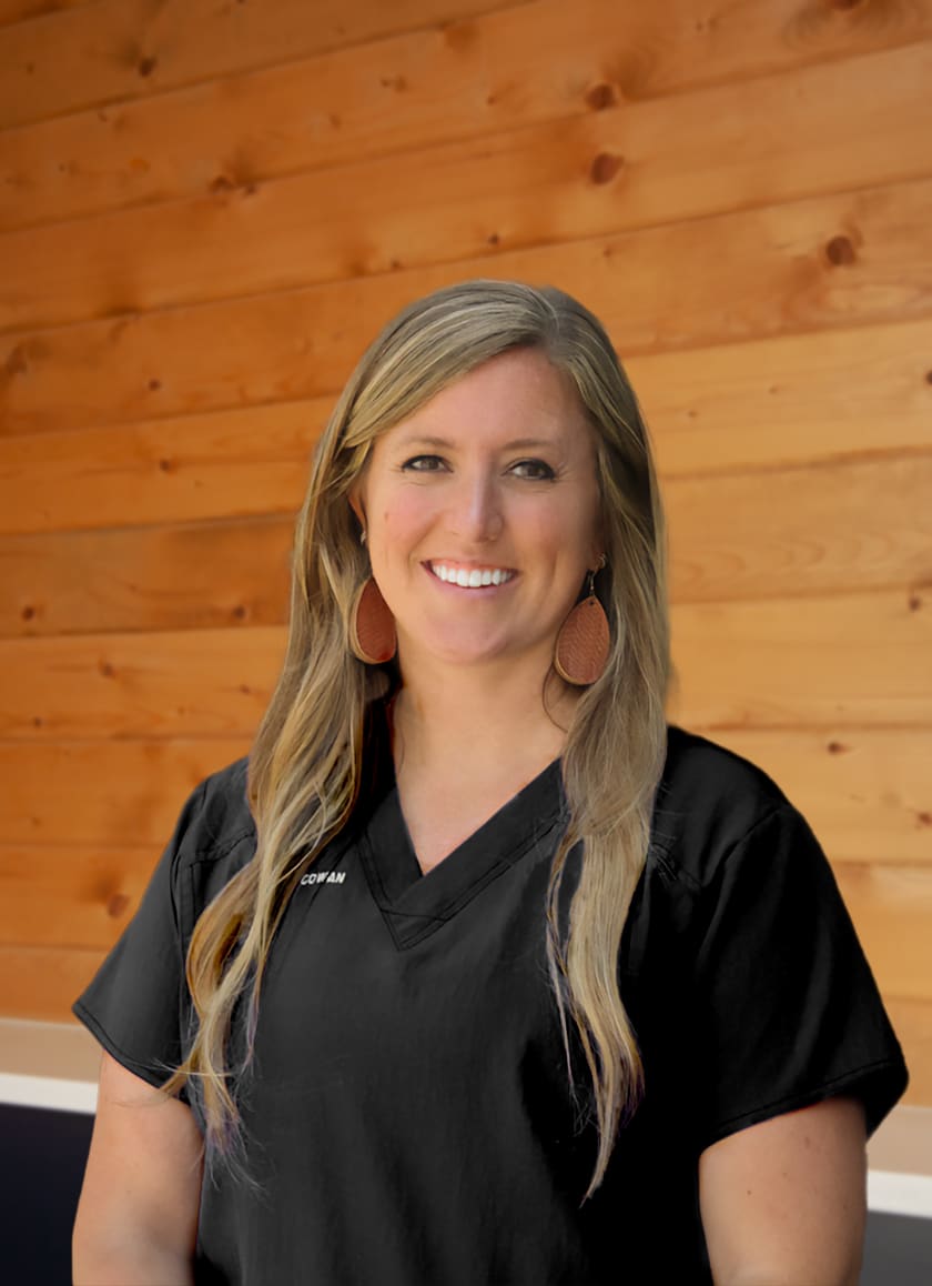 A smiling woman with long blonde hair wearing black medical scrubs and large brown earrings stands in front of a wooden wall.