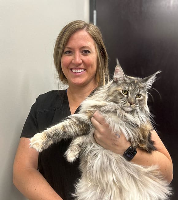 A woman with shoulder-length blonde hair, wearing a black shirt and a smartwatch, smiles while holding a large, fluffy tabby cat with white and grey fur. As a dedicated veterinarian, she stands indoors against a light-colored wall with a dark door in the background.